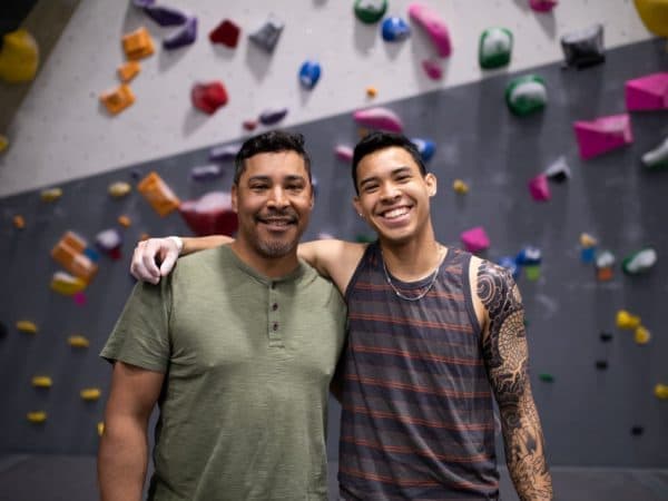 Two smiling men stand with arms around each other in front of a climbing wall