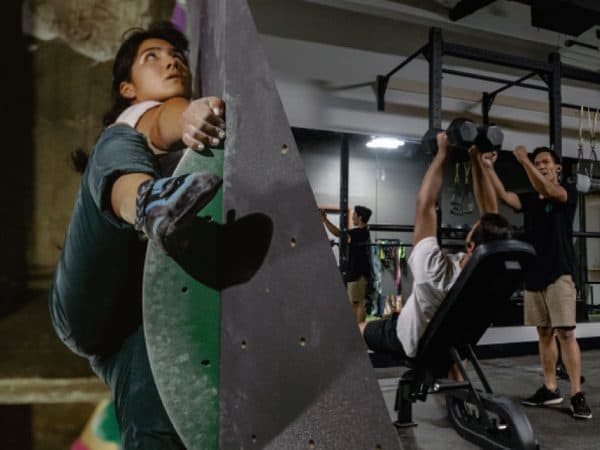 A woman climbs a green-edged wall while people lift dumbbells on a bench in the fitness area behind