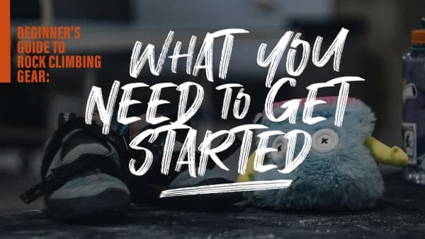 Pair of climbing shoes next to a fuzzy blue chalk bag and a water bottle on a dusty floor