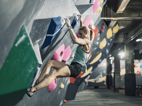 Blonde woman in a teal tank top swings across a colorful steep bouldering wall inside a warehouse-style gym