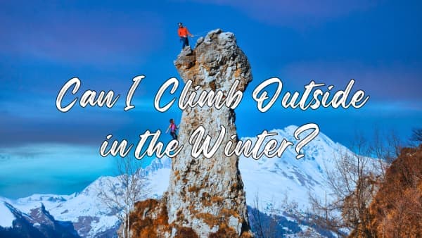 Two climbers in bright jackets stand on a tall rock spire in snowy winter mountains under a blue sky