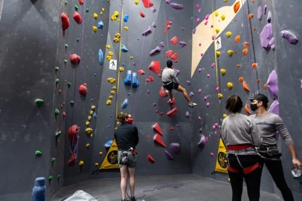 Climber scales a tall indoor wall while two harnessed belayers in masks and another person watch from below
