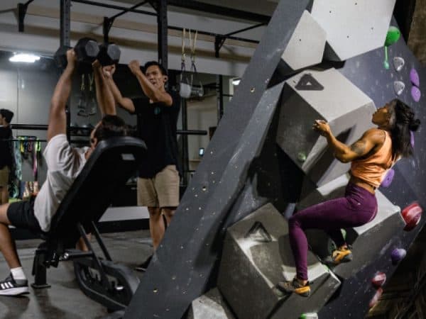 Woman climbs a gray angular bouldering wall while people lift dumbbells on a bench in a gym behind her