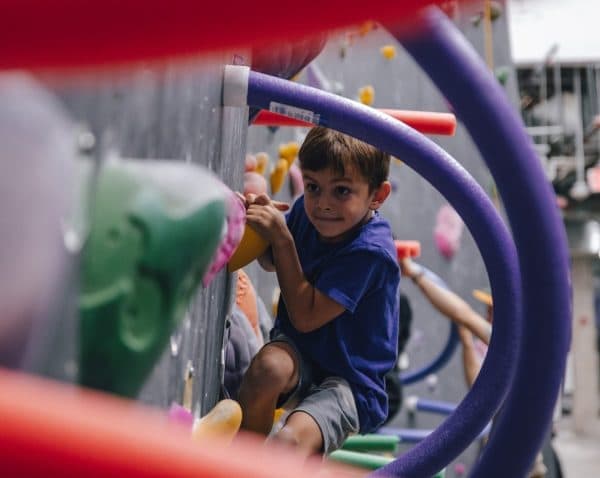 Young boy in a blue shirt climbs a bouldering wall, seen through blurry foam pool noodles and colorful holds