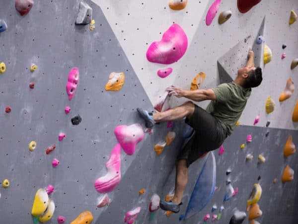 A man in a green shirt climbs a gray wall with pink and orange holds, one foot braced