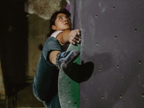 A climber with dark hair looks upward while gripping a green hold and bracing a foot on the wall