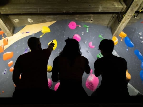 Three people in silhouette sit and point at a climbing wall with pink, yellow, orange and blue holds