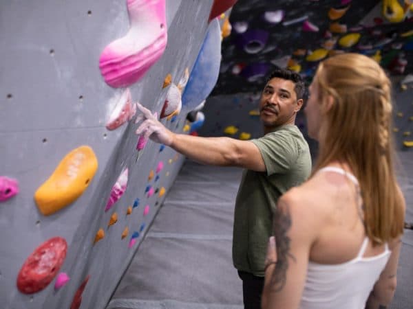 A climbing coach in a green shirt points out a hold to a woman with a braided ponytail