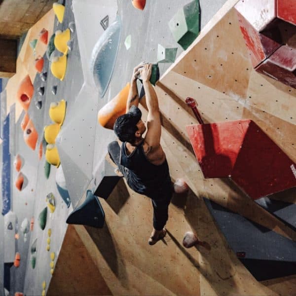 A man in a black tank top hangs from two handholds on a tall bouldering wall of colorful holds