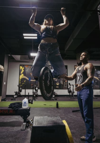 Woman in a cap does a weighted pull-up with a plate on her belt while a coach spots her