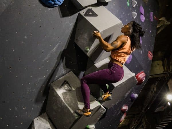 A woman in an orange top and purple leggings climbs a dark bouldering wall with gray angular volumes