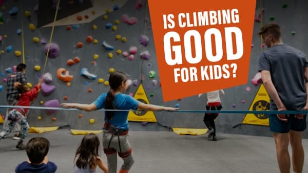 Young girl in a harness balances with arms outstretched while other kids and an adult play near a colorful climbing wall