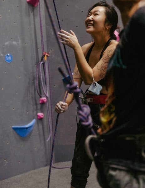 Smiling climber in a harness stands beside a rope at an indoor wall, laughing while looking up at a partner