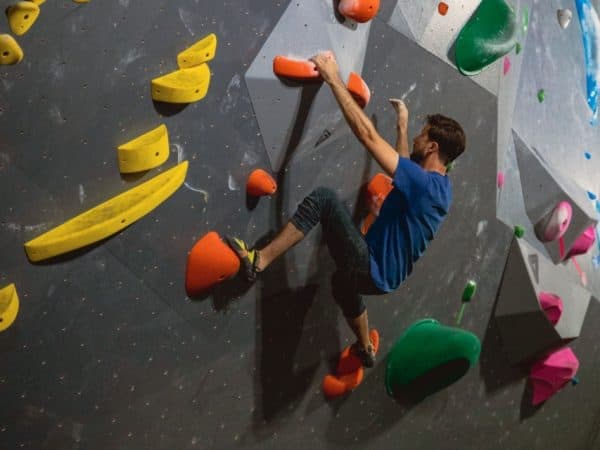 A climber in a blue shirt stretches up a dark bouldering wall studded with orange and yellow holds