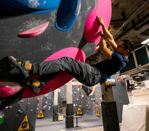 Climber in glasses hangs from pink holds on a steep indoor bouldering overhang, with a gym behind him