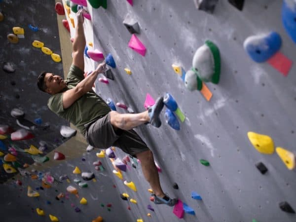 A man in an olive shirt climbs a gray wall at a steep angle, gripping a handhold with chalked fingers