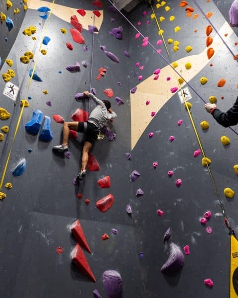 Climber ascends a tall indoor wall covered in colorful holds on a top rope while a belayer holds the line