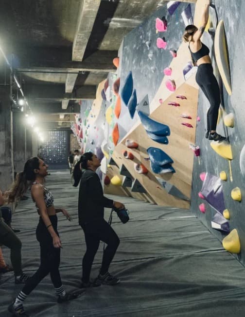Climber on a tall colorful wall while others watch from the mats