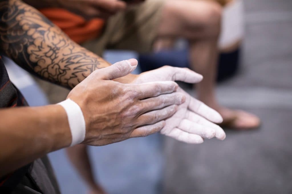 Close-up of a tattooed climber rubbing white chalk onto both hands, with other people blurred in the background