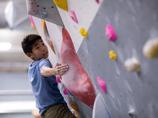 A young boy in a blue shirt reaches for a large red hold on an indoor climbing wall