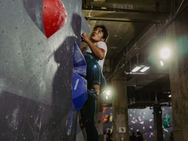 A climber in a light shirt looks up while pressing a foot on a blue hold, concrete ceiling above