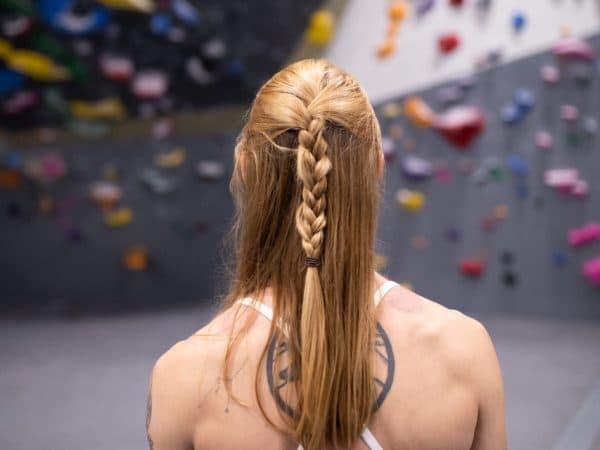 A woman with a long braided ponytail faces a bouldering wall, seen from behind in a climbing gym
