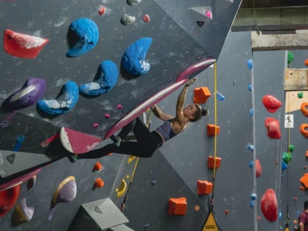 A woman in a sports top reaches overhead on a steep climbing wall with blue and red holds